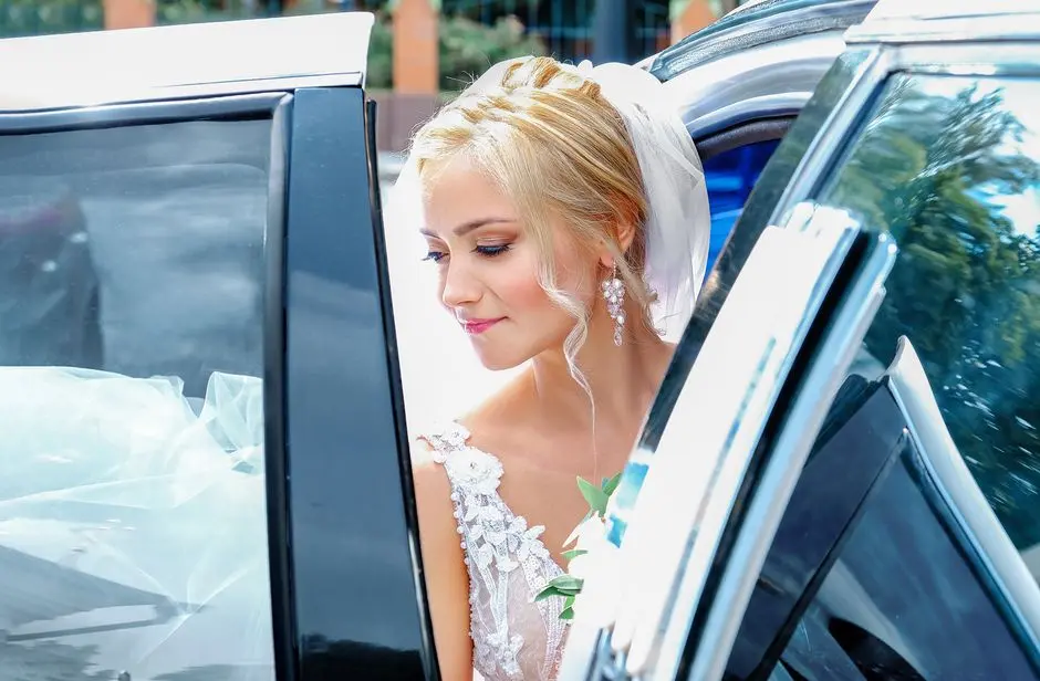 Bride med elegant oppstilt blondt hår og slør stiger ut av bryllupsbil, eksempel på brudehår 2026.