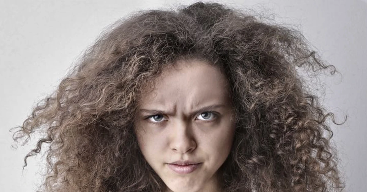 Frizz hår: Kvinne med krøllete, utslettet hår og skeptisk ansiktsuttrykk mot grå bakgrunn.